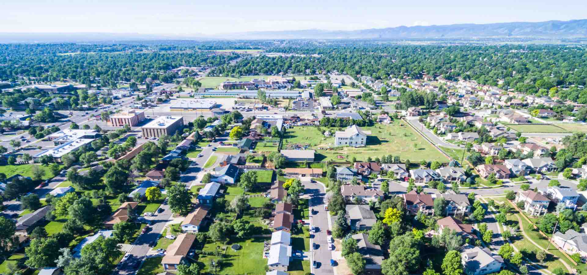 Aerial view of a suburban neighborhood with tree-lined streets, houses, and buildings, set against a backdrop of distant mountains and clear skies.