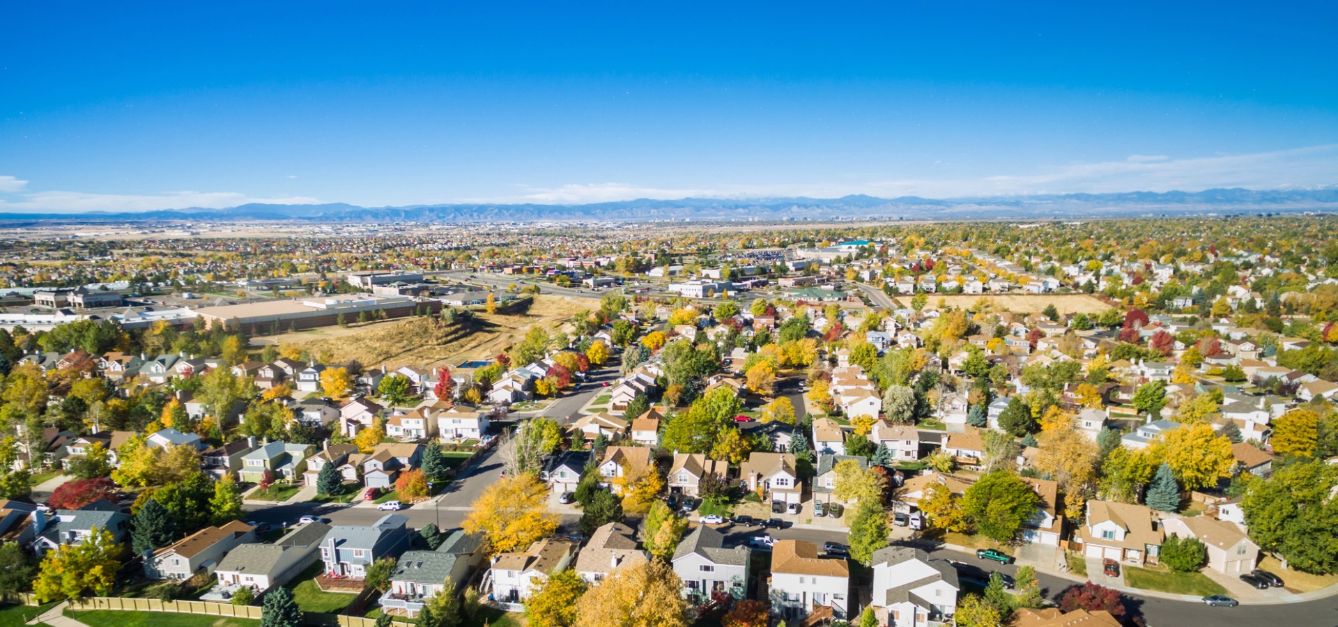 Aerial view of a suburban neighborhood in Denver, Colorado, with tree-lined streets, colorful autumn foliage, and houses, set against a backdrop of distant mountains and a clear blue sky.