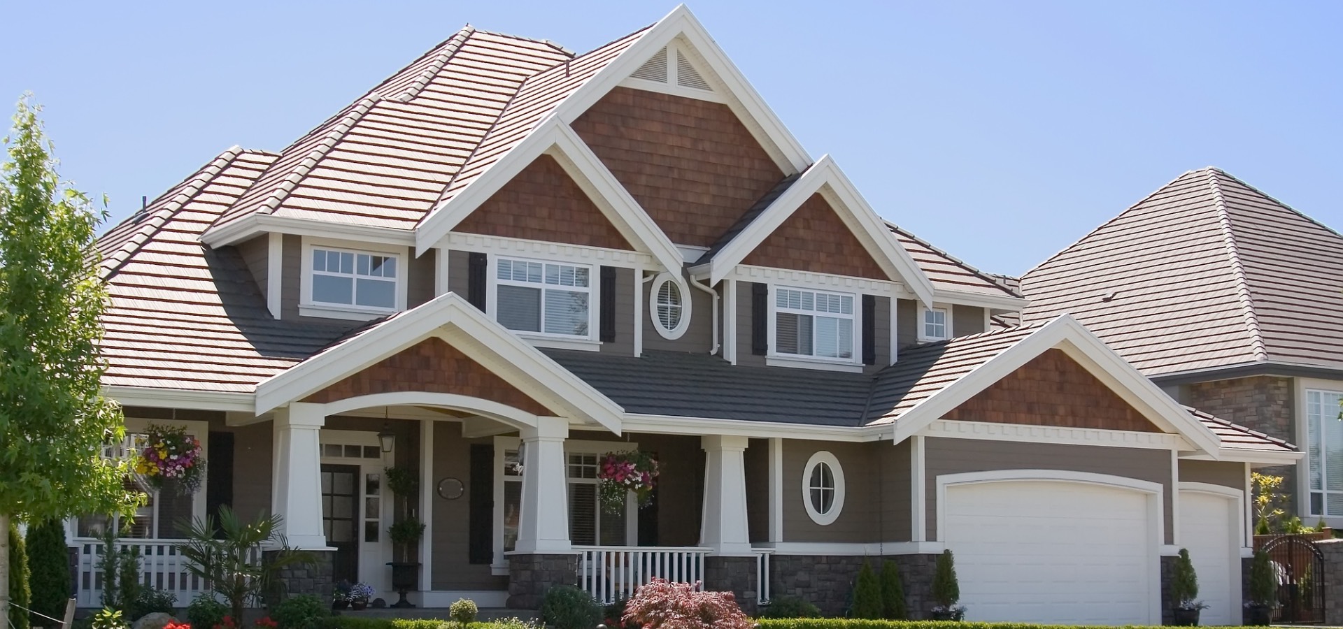 A large, modern suburban home with a brown and white exterior, featuring multiple gables, a covered front porch, and a well-manicured lawn, located in a residential neighborhood.